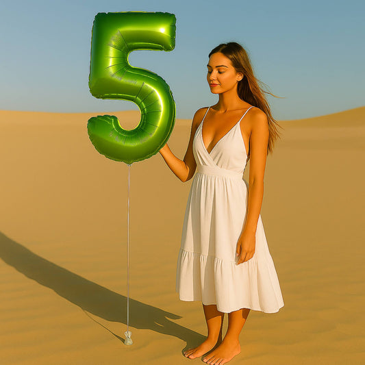 Woman in a white dress holding a large green balloon shaped like the number five in a desert setting.