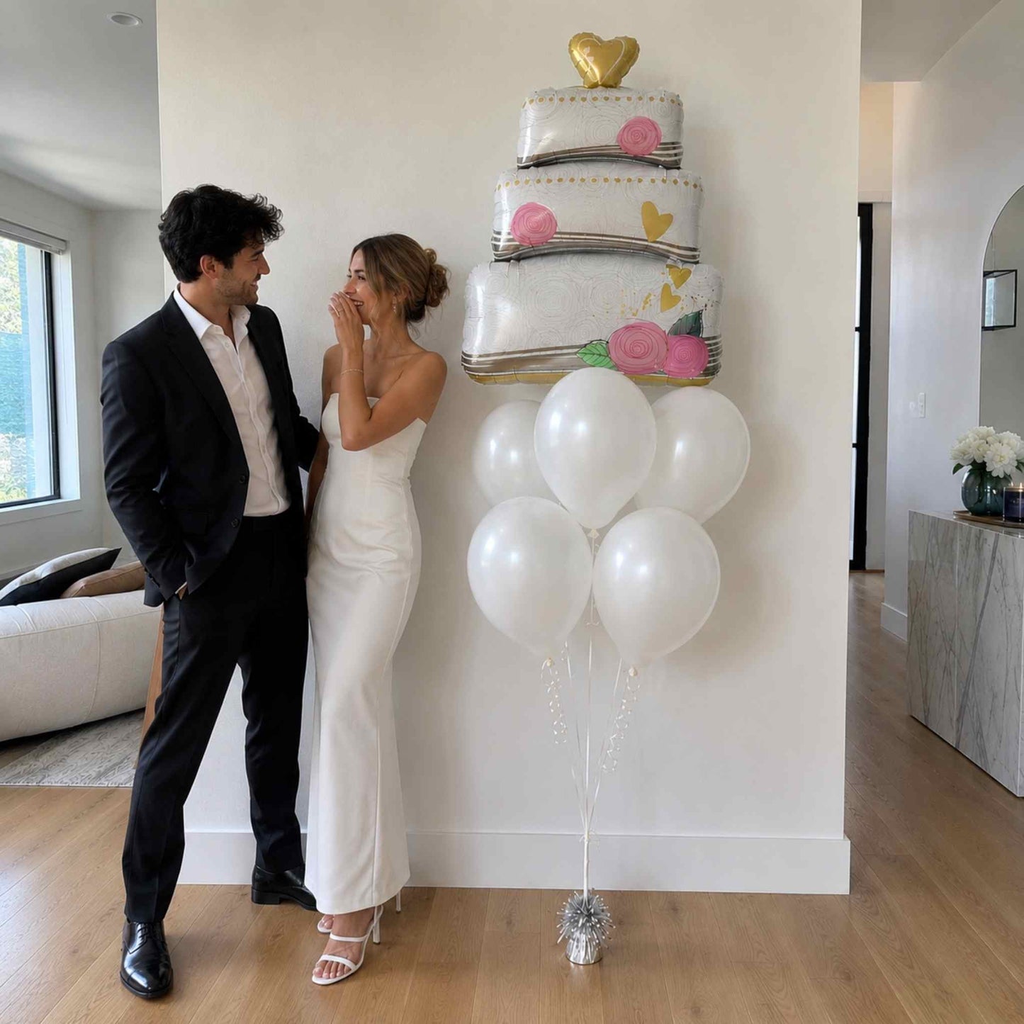 A couple in formal wedding attire stands indoors beside a decorative wall arrangement featuring a tiered cake design and a cluster of white balloons.