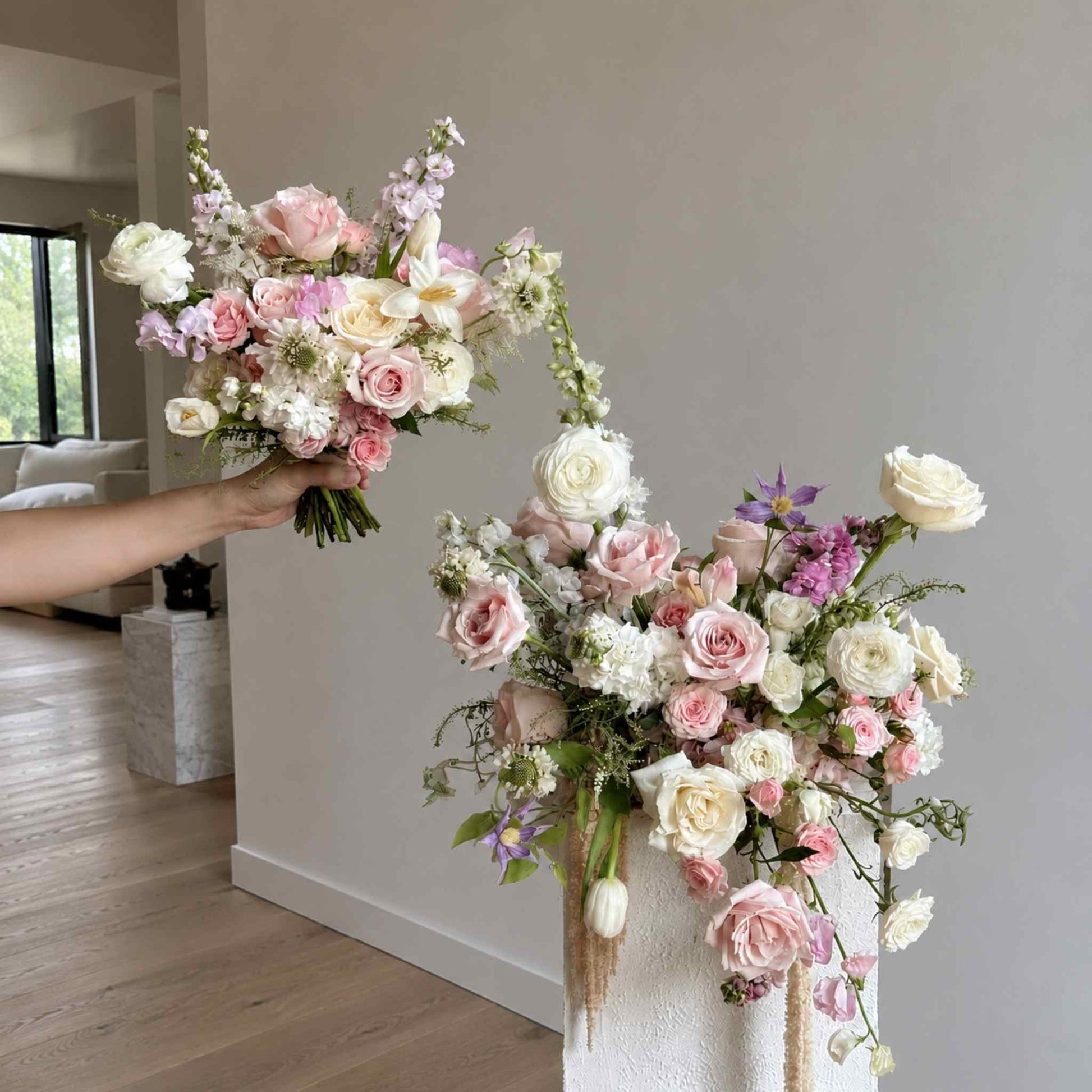 Person holding a bouquet of flowers next to a large floral arrangement indoors.