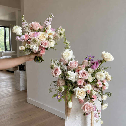 Person holding a bouquet of flowers next to a large floral arrangement indoors.
