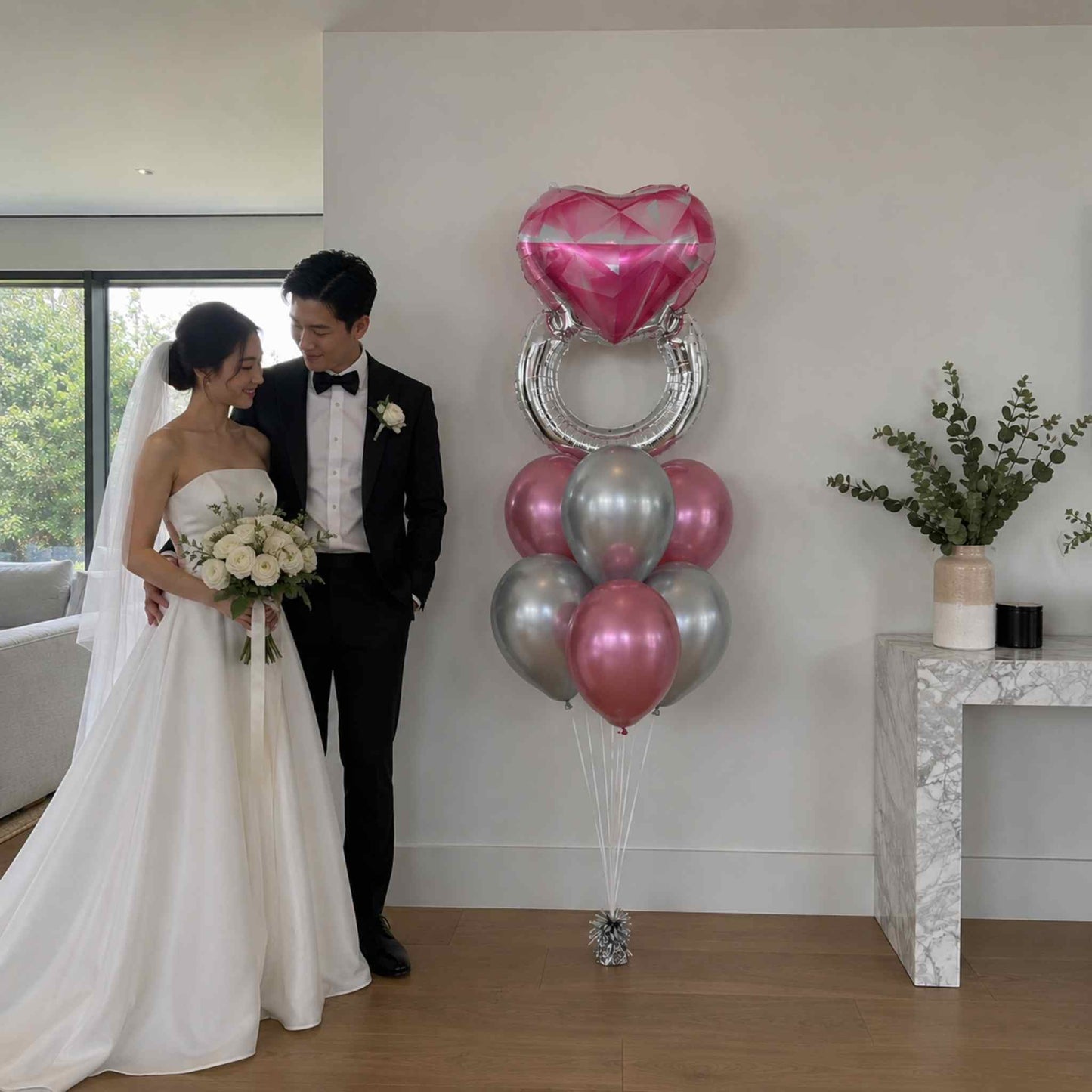 Wedding couple standing next to a decorative arrangement of pink and silver balloons in a room with a window and plant.