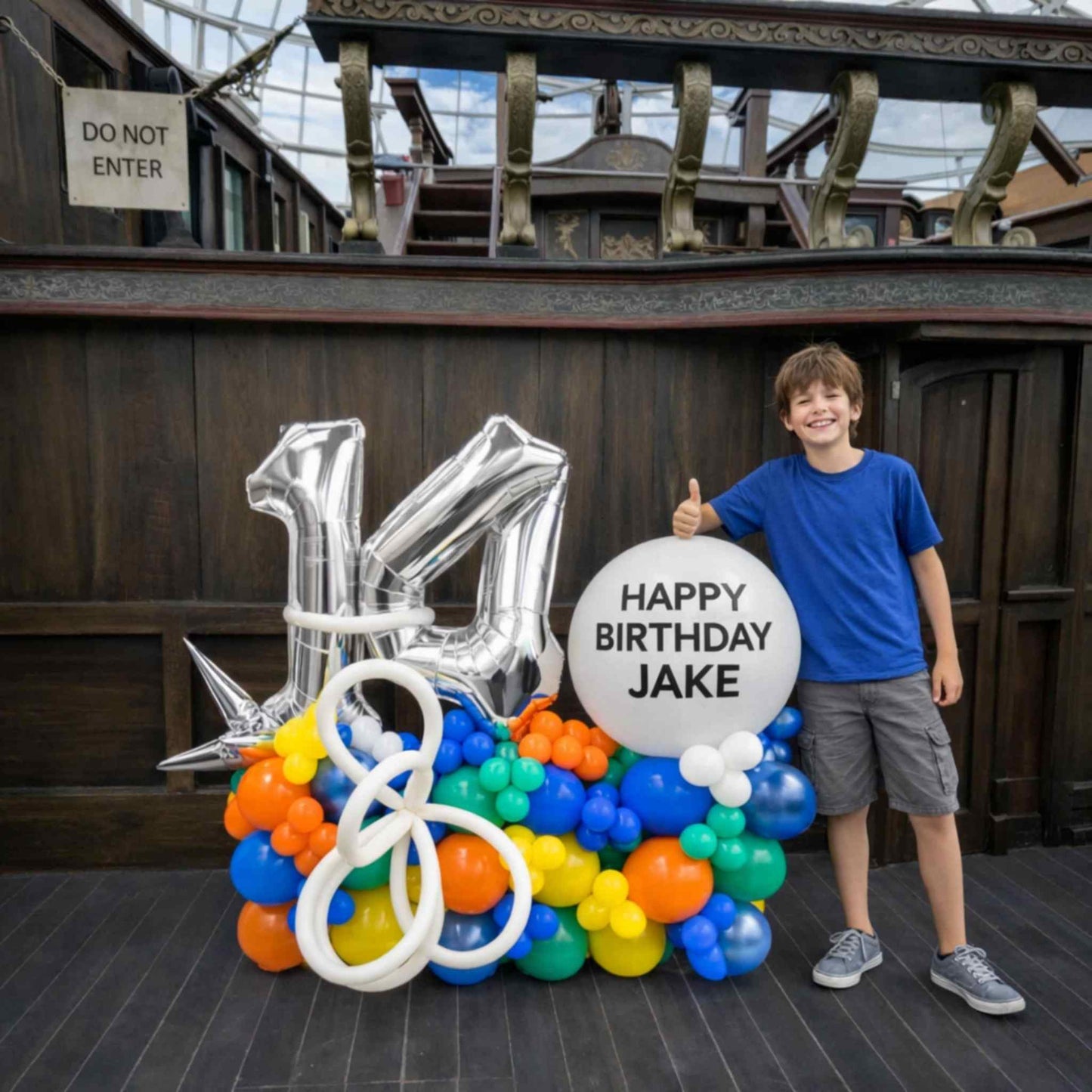 Child standing next to a birthday balloon arrangement with 'Happy Birthday Jake' sign in front of a wooden wall.