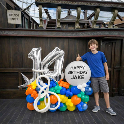 Child standing next to a birthday balloon arrangement with 'Happy Birthday Jake' sign in front of a wooden wall.