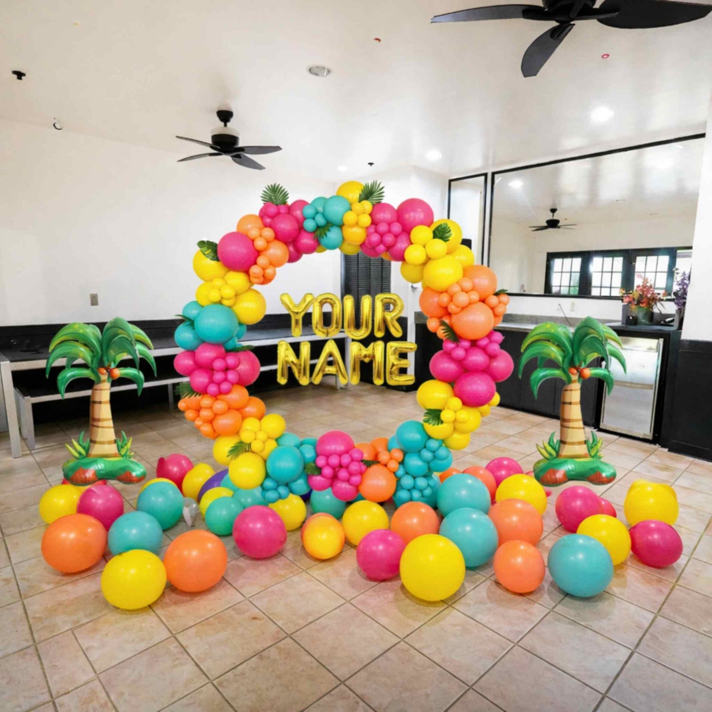 Colorful balloon arch with 'YOUR NAME' text in a room with palm trees and a ceiling fan.