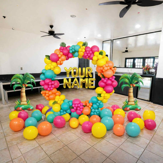 Colorful balloon arch with 'YOUR NAME' text in a room with palm trees and a ceiling fan.