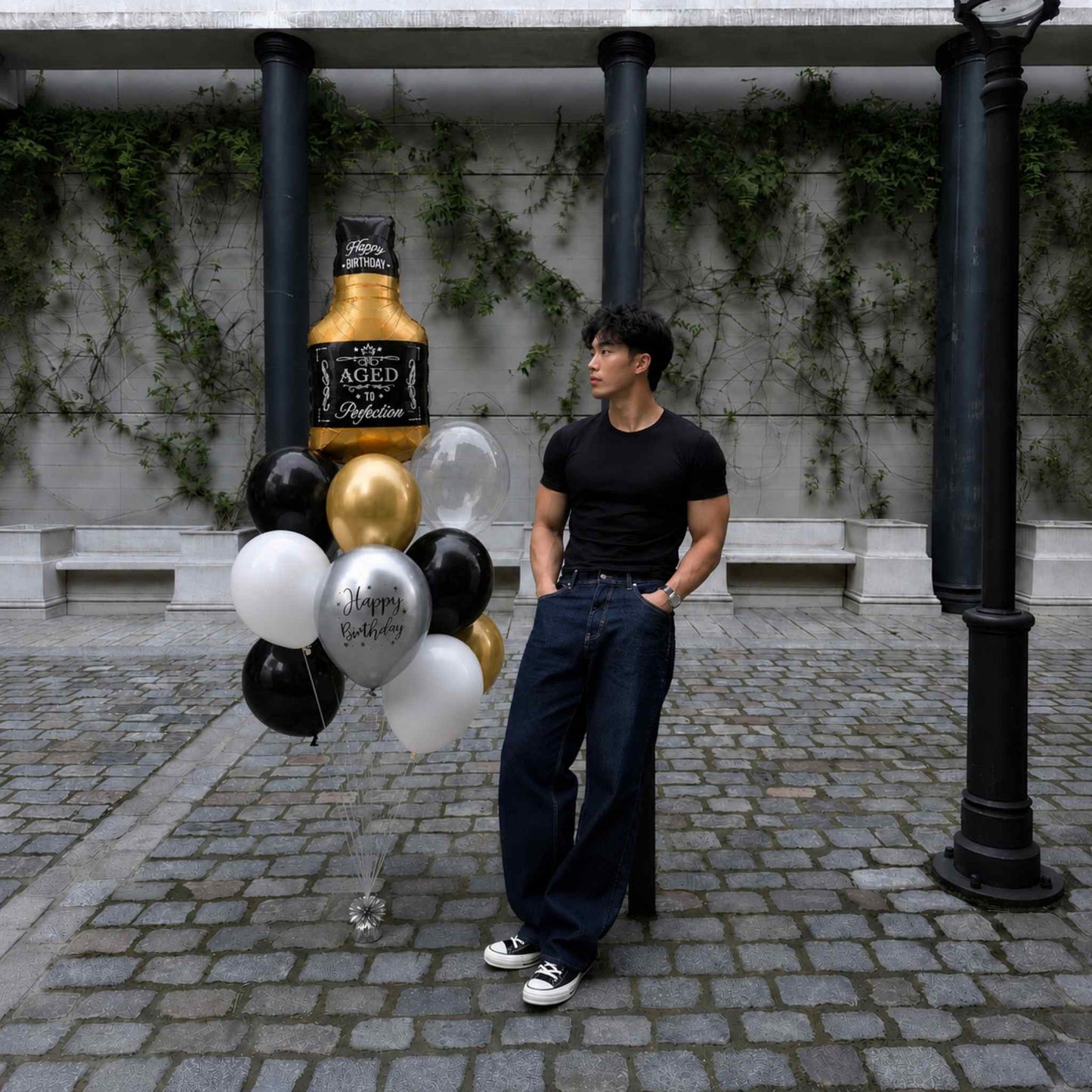 A man in a black t-shirt and dark jeans stands outdoors next to a cluster of black, white, and gold balloons, including a large balloon shaped like an aged whiskey bottle labeled 'Aged 15 AgED'.