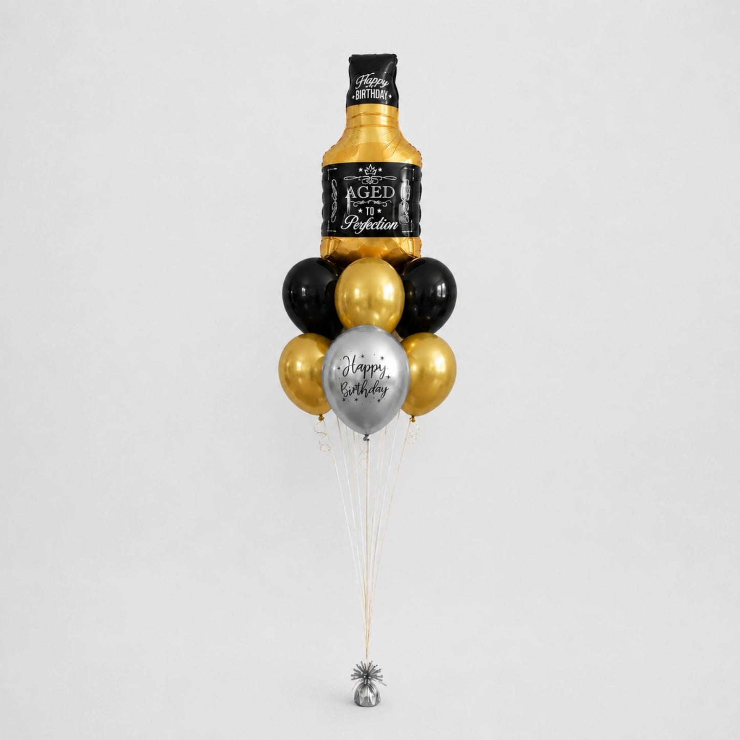 A novelty balloon bouquet designed to look like a bottle of aged whiskey, with black and metallic gold balloons, a silver balloon reading 'Happy Birthday', and a small silver bell at the bottom, photographed on a light gray background.