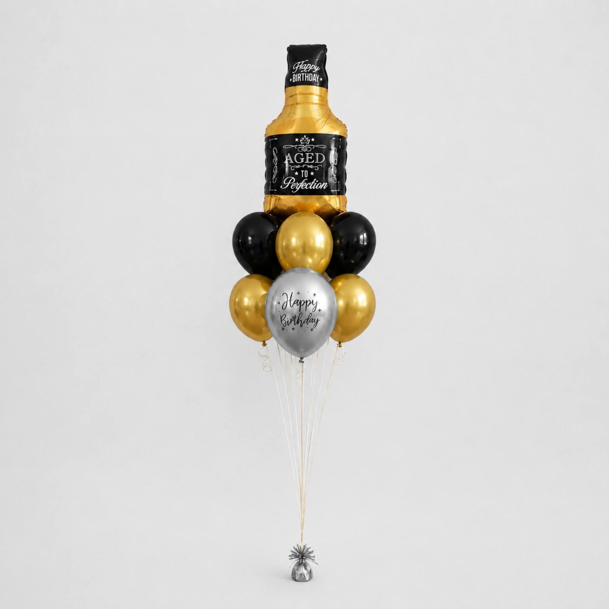 A novelty balloon bouquet designed to look like a bottle of aged whiskey, with black and metallic gold balloons, a silver balloon reading 'Happy Birthday', and a small silver bell at the bottom, photographed on a light gray background.