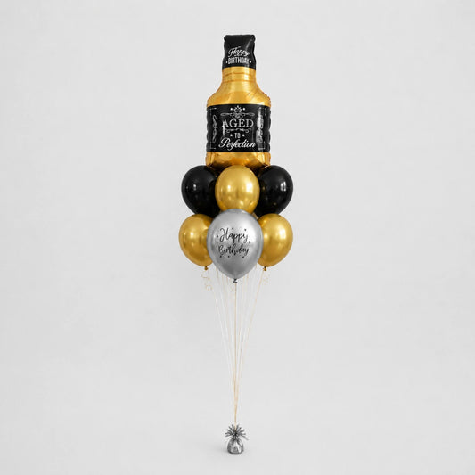 A novelty balloon bouquet designed to look like a bottle of aged whiskey, with black and metallic gold balloons, a silver balloon reading 'Happy Birthday', and a small silver bell at the bottom, photographed on a light gray background.