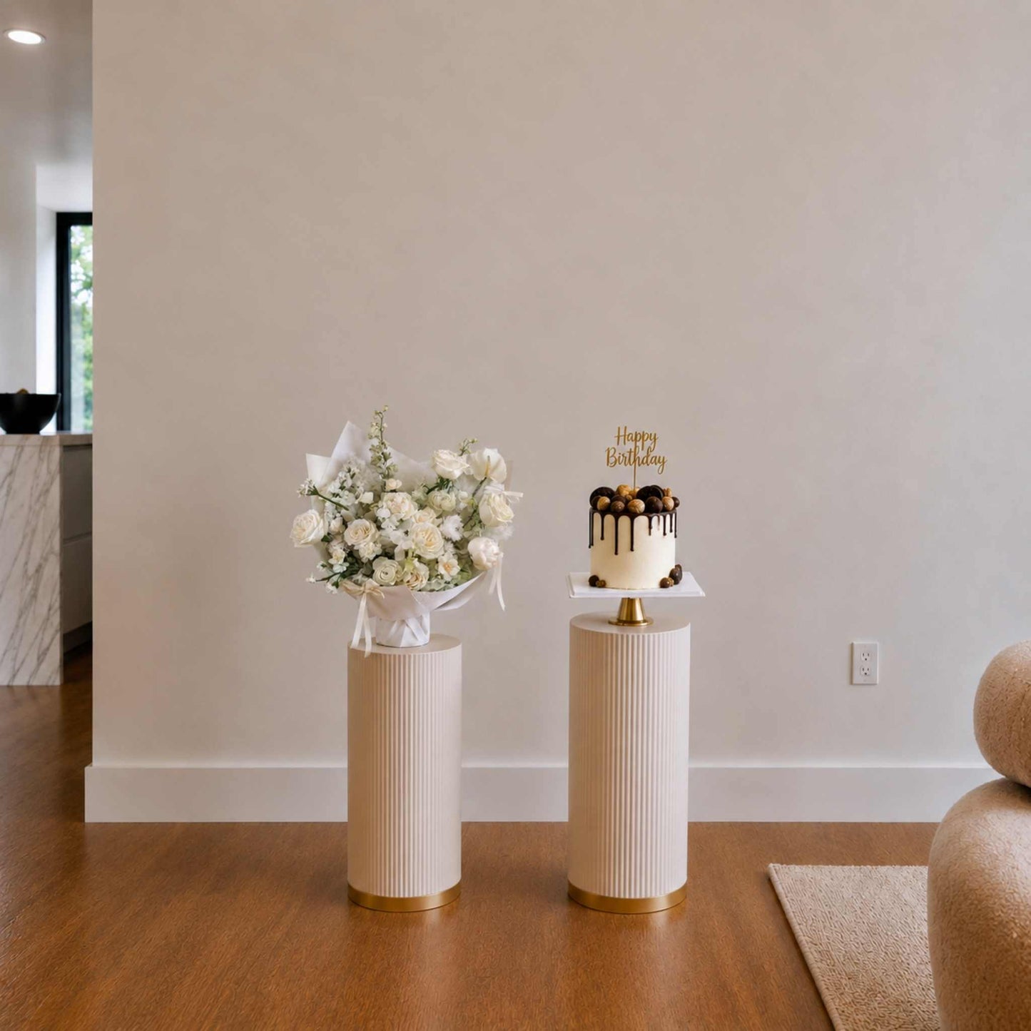 Decorative cake and floral arrangement on white stands against a plain wall.