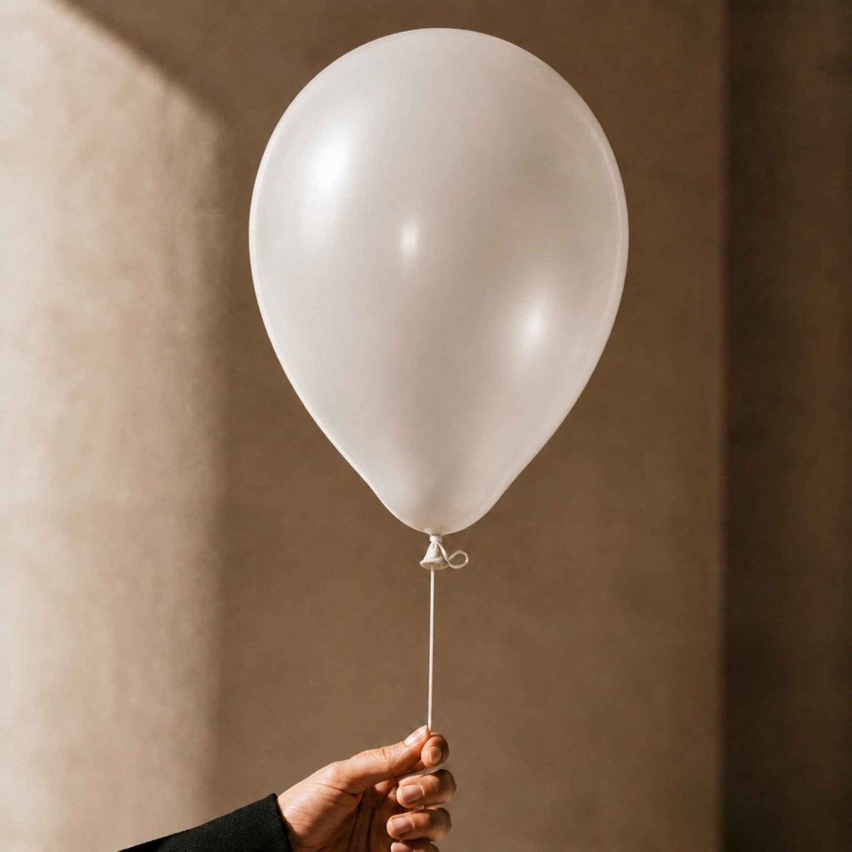A single matte white balloon held by a hand against a warm beige textured wall.