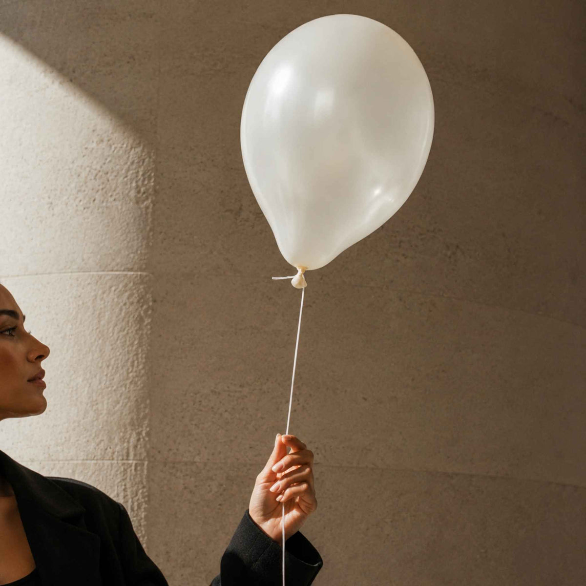 A person in a black blazer holds a translucent white balloon against a textured beige wall with dramatic sunlight and shadow.