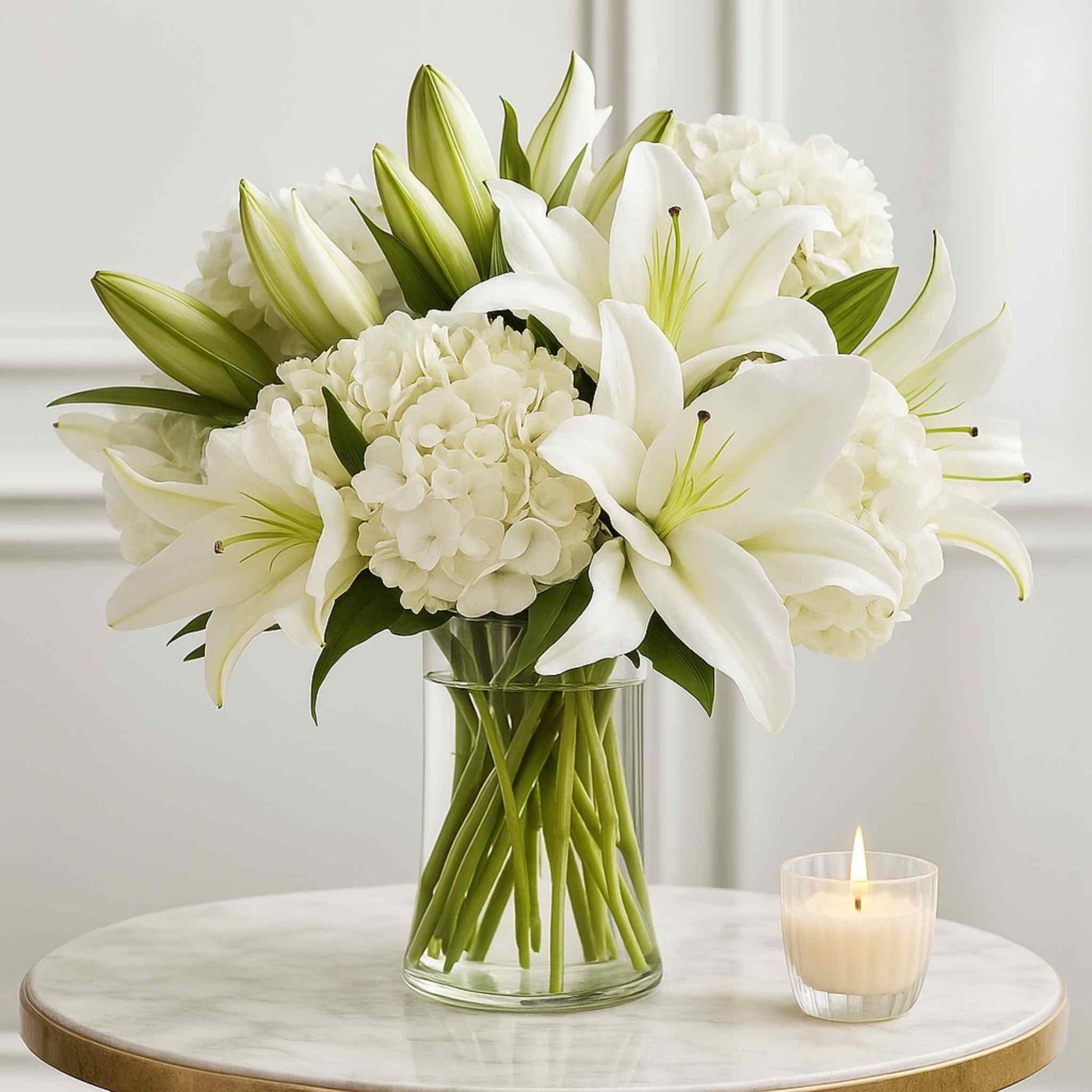 Bouquet of white lilies and hydrangeas in a clear vase on a marble table with a lit candle.