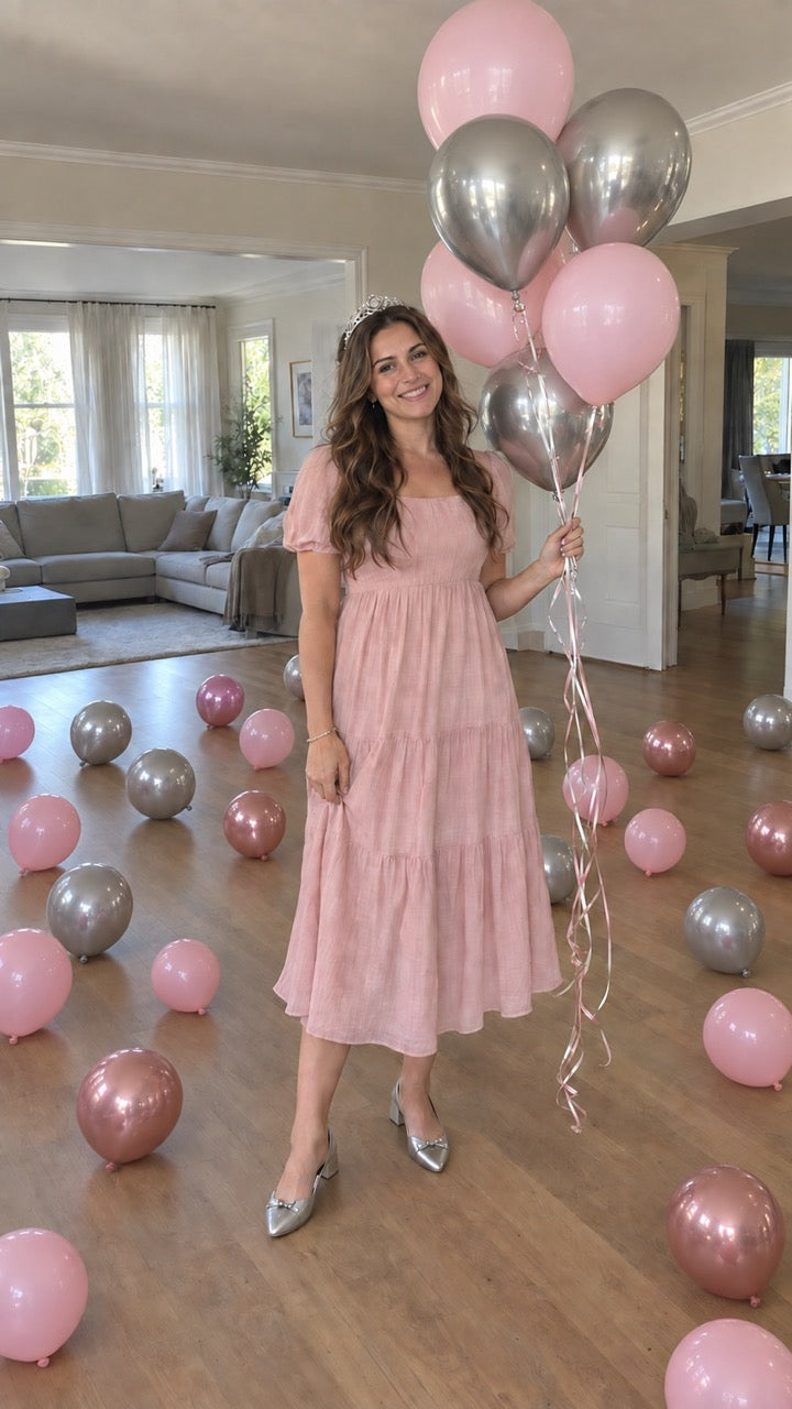 A woman stands in a bright living room holding a cluster of pink and metallic silver balloons. She is wearing a light pink tiered midi dress and metallic silver heels.