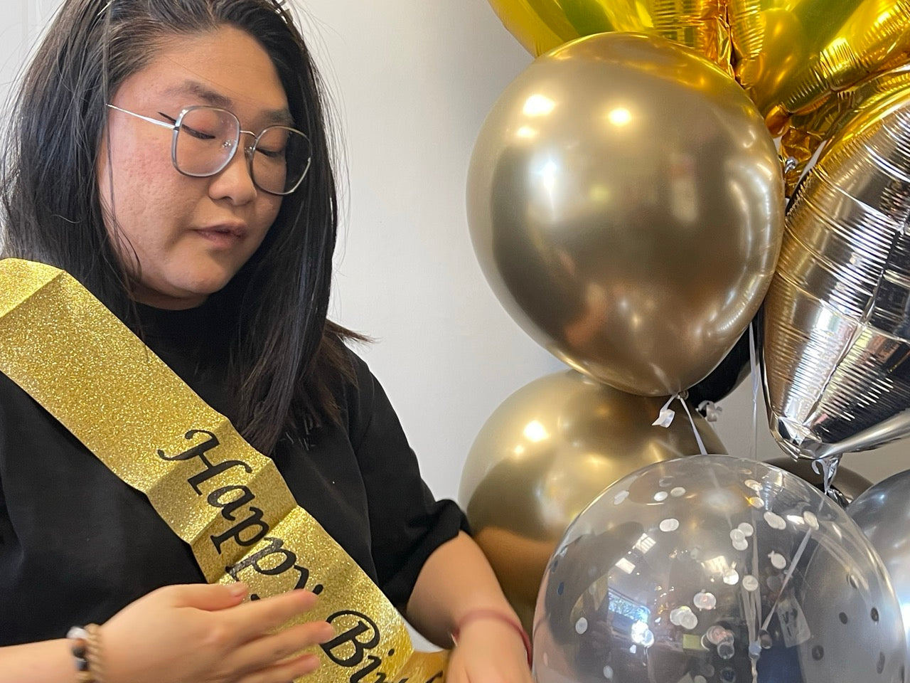 A woman wearing glasses and a black top is shown from the side, wearing a glittery gold 'Happy Birthday' sash. She is standing next to a cluster of metallic gold, silver, and clear balloons with confetti.