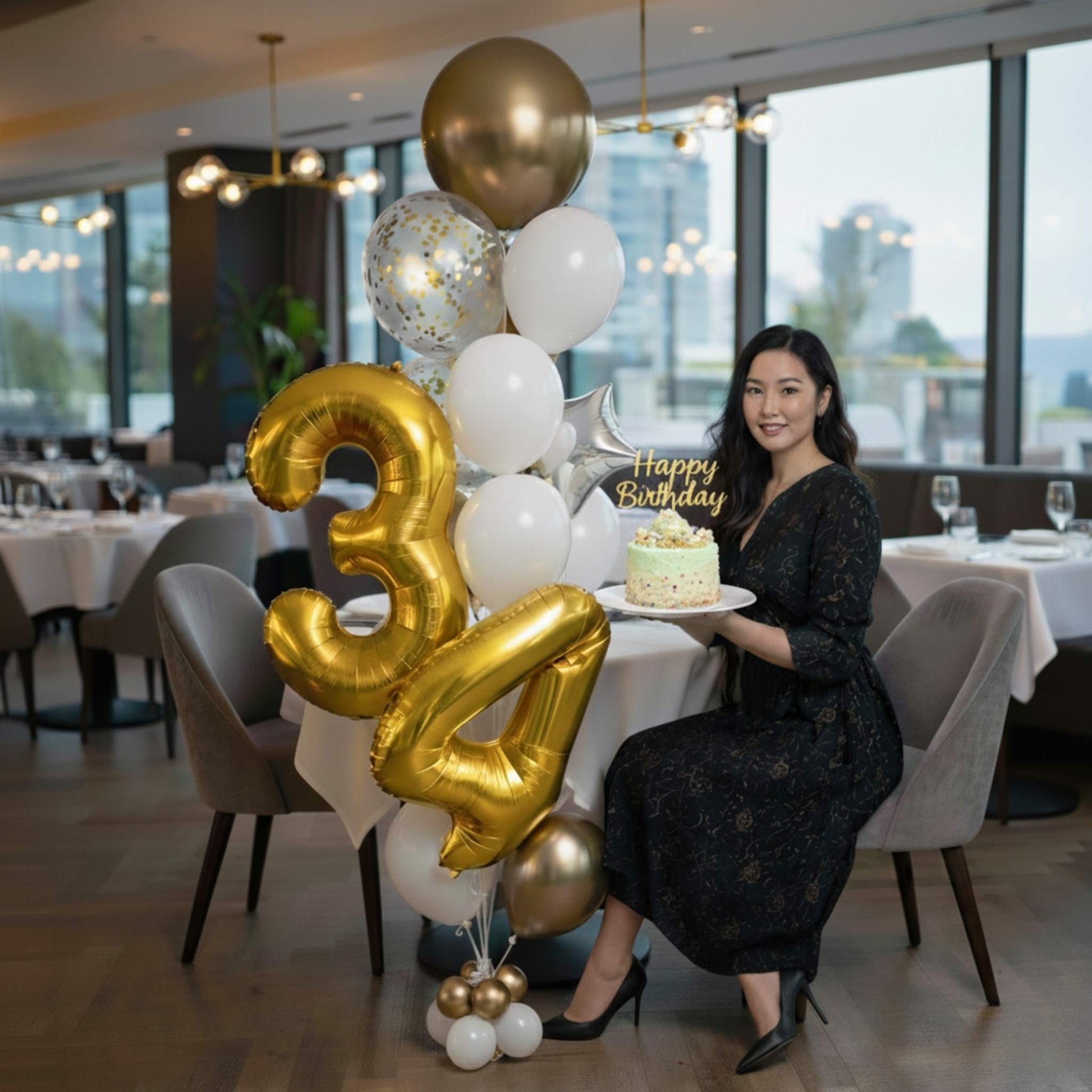 Woman holding a birthday cake with gold and white balloons in a restaurant setting.
