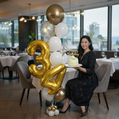 Woman holding a birthday cake with gold and white balloons in a restaurant setting.