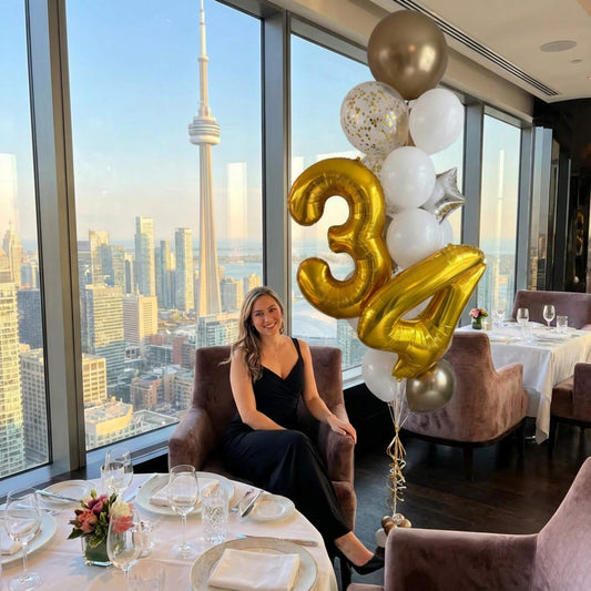 Woman sitting in a chair with large '34' balloons in a room with a cityscape view.
