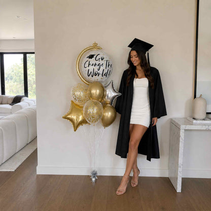 Graduate in cap and gown standing next to balloons with a motivational message in a living room.