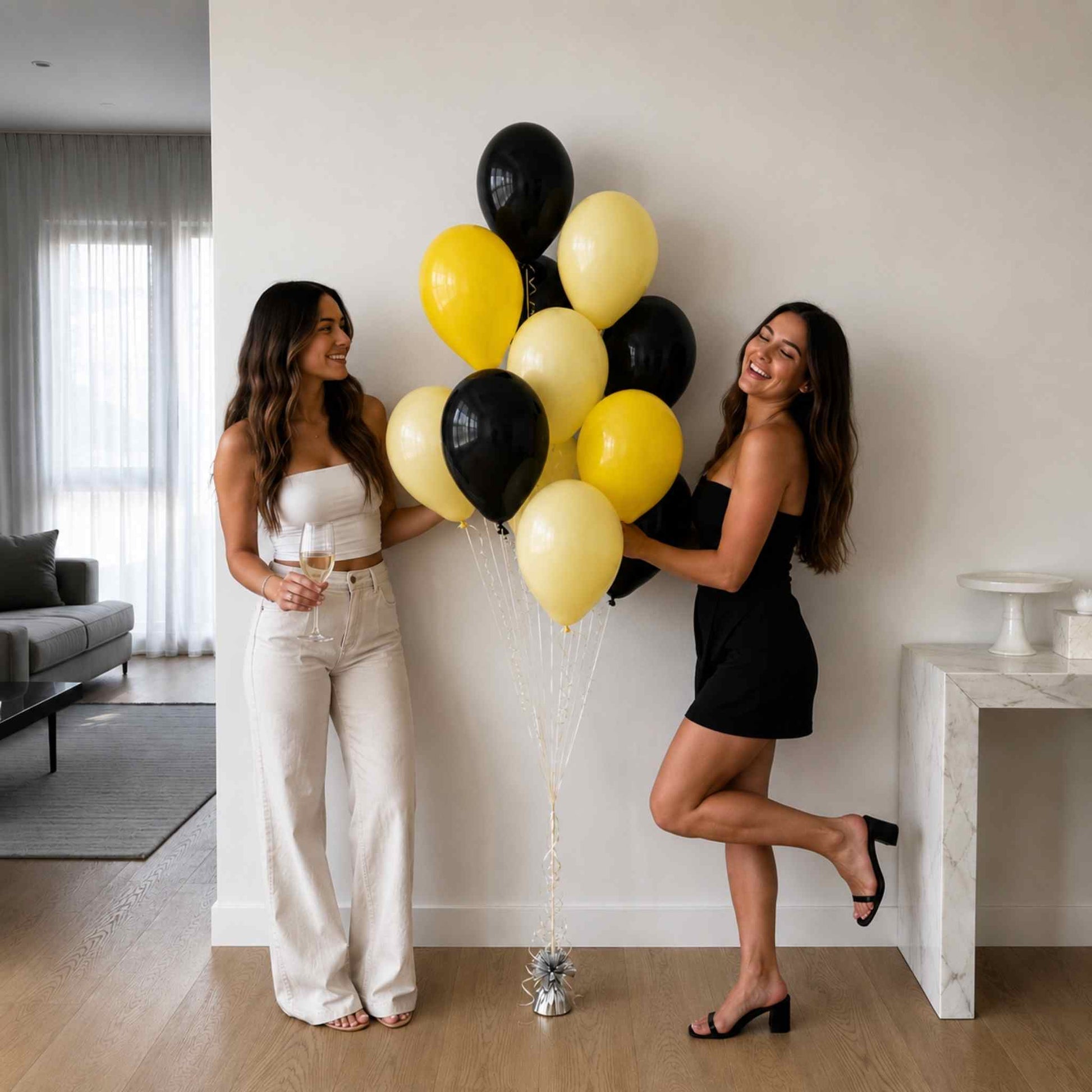 Two women standing in a bright living room holding a cluster of black and yellow balloons. One woman is wearing a white strapless top and white wide-leg pants, and the other is wearing a black strapless mini dress and black heels.