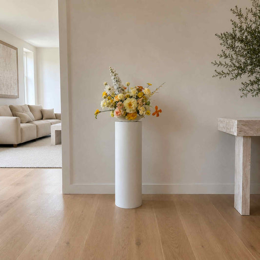 Floral arrangement in a white vase on a wooden floor with a neutral-colored wall and furniture in the background.