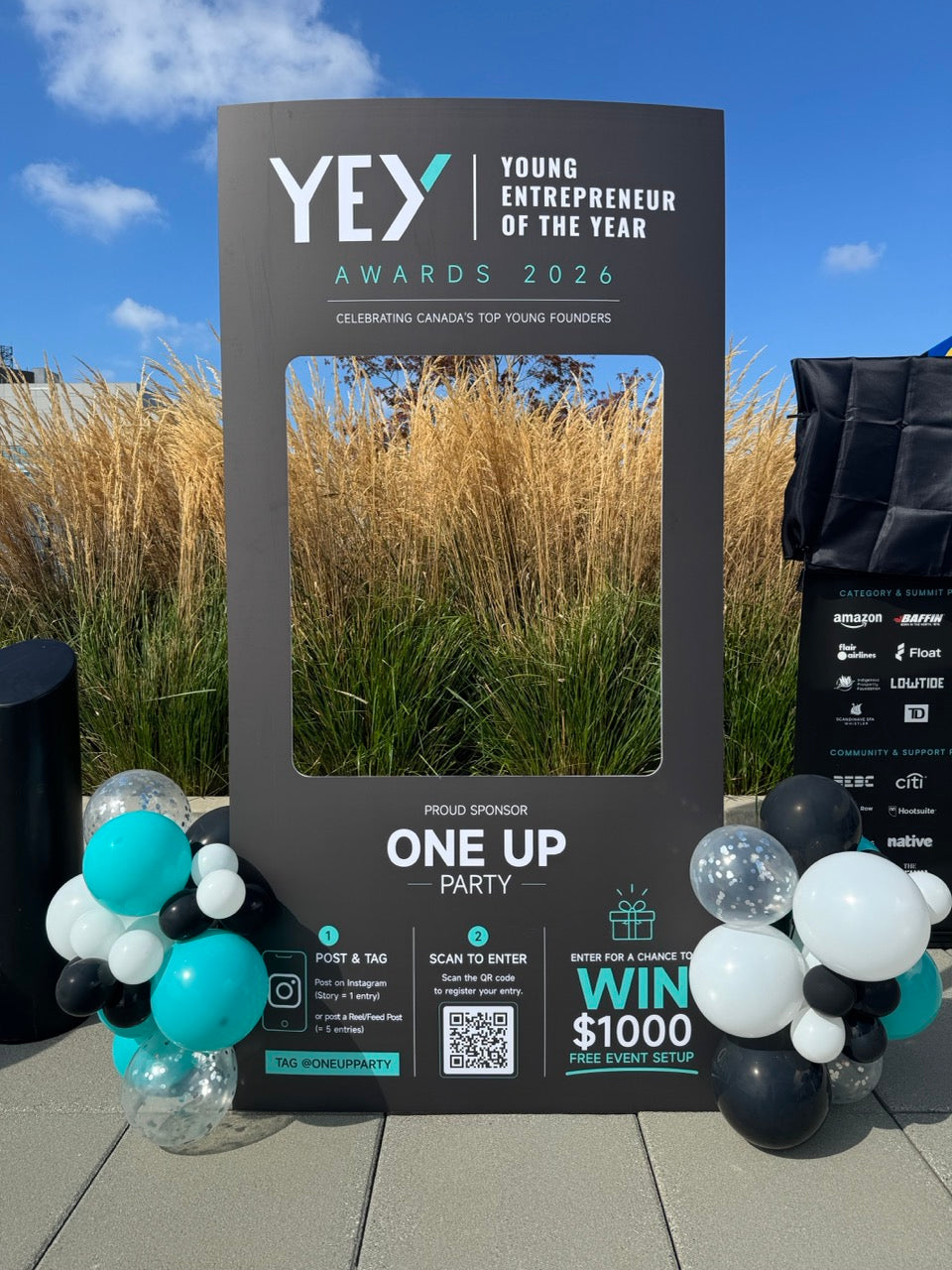 A large black photo booth frame stand for the YEW Young Entrepreneur of the Year Awards 2026, set outdoors on a tiled patio with decorative teal, black, and white balloon clusters on either side. The backdrop features dry golden grass and a blue sky.